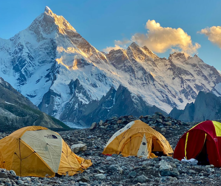 Tents at a high-altitude campsite with a breathtaking view of the Karakoram mountains, including a snow-covered peak glowing under the golden sunset.